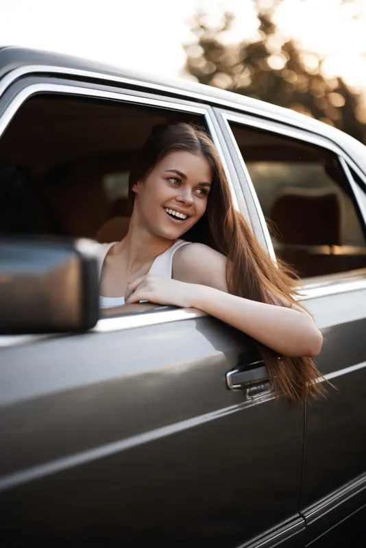 Young woman with long hair joyfully leaning out of a car window, enjoying a sunny day, capturing the essence of freedom and adventure.
