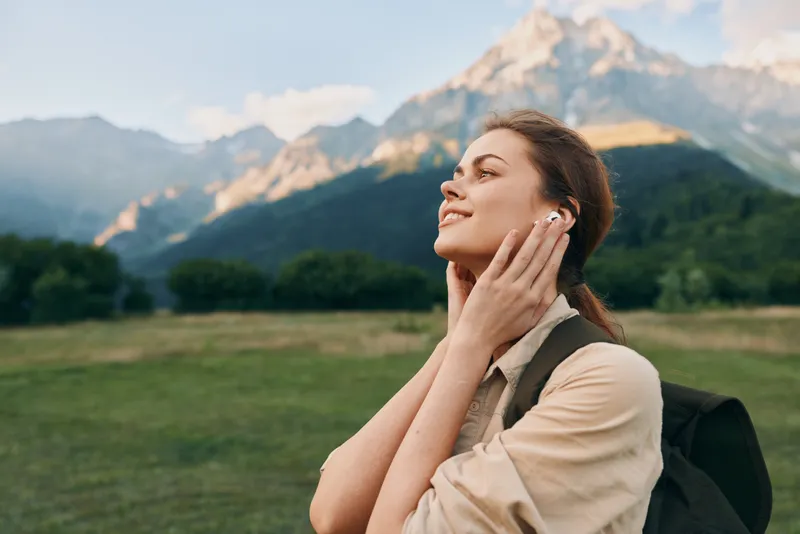Smiling woman outdoors enjoying nature with a mountain backdrop and grassy field, wearing a light backpack, embodying a relaxed lifestyle and wellness moment.