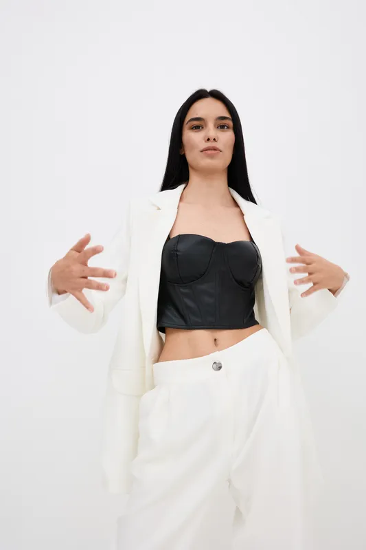 Young woman wearing white suit and black corset top posing confidently against plain white background, showcasing modern casual style and minimalistic fashion.