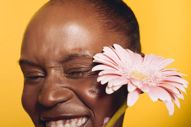 Close-up of a happy black woman with dark skin holding a pink gerbera flower near her face against a yellow background. Concept of natural beauty, joy, and floral skincare.