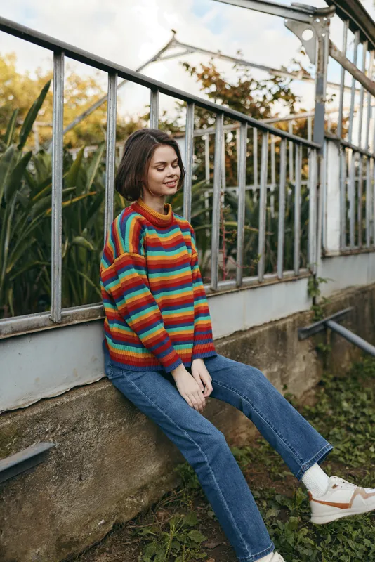 woman sweater jeans greenhouse sitting casual young woman in colorful striped knit sweater and blue jeans relaxing by a metal greenhouse railing among plants and sunlight
