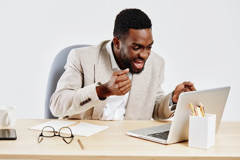 Happy young African American man wearing beige suit jacket working excitedly on laptop at desk in bright office. Business casual, success, communication, remote work, smiling, people concept, modern w