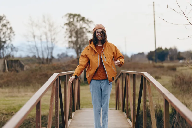 Confident woman lifestyle portrait, walking on a wooden bridge outdoors in autumn, orange jacket and beanie, calm rural park background, soft natural light, candid.