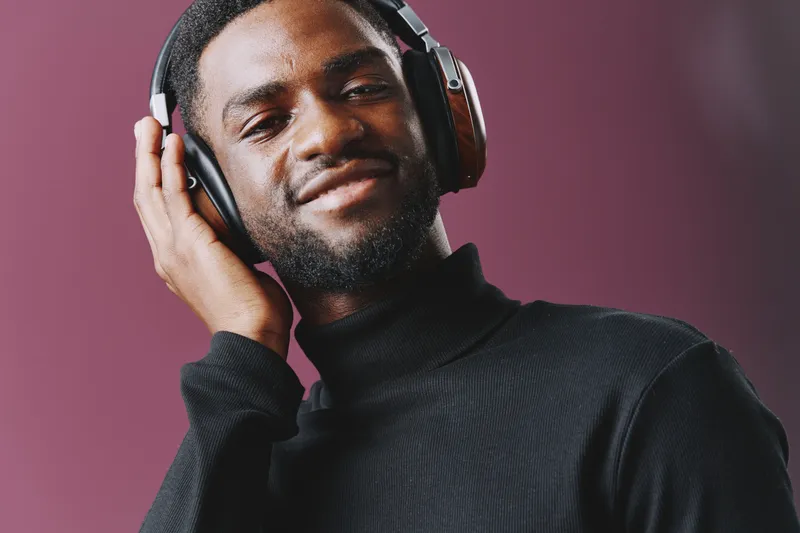 Young Black man enjoying music with headphones, wearing a stylish black turtleneck sweater, smiling and relaxed against a purple background