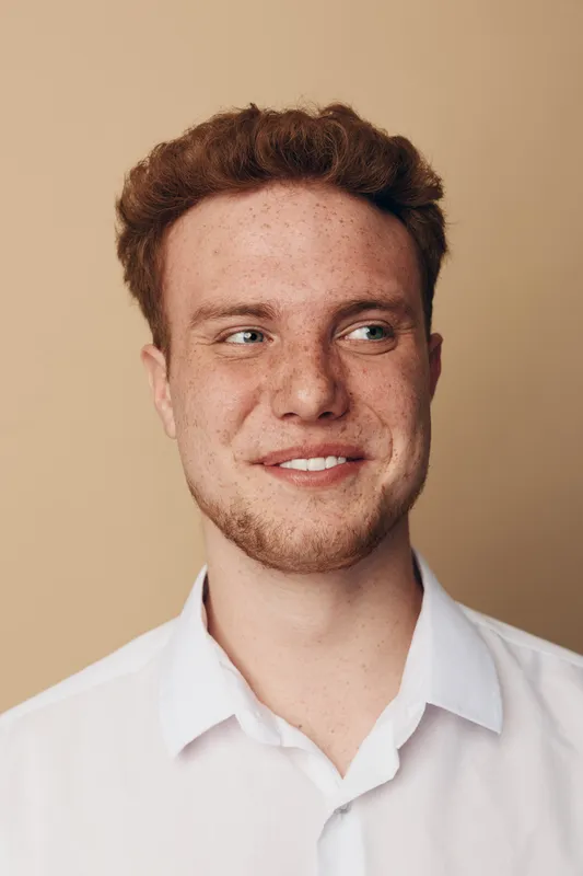 Young man with red hair and freckles smiles gently looking sideways. Casual portrait on beige background with natural light. Concept of youth, happiness, and natural beauty.
