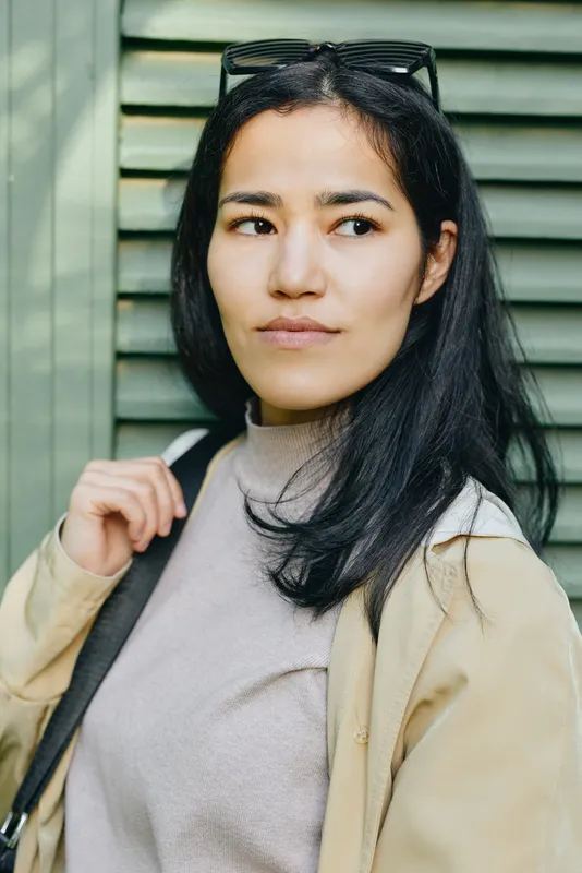 Woman in beige coat and light sweater outdoors with sunglasses on head, thoughtful expression, urban backdrop of green slats and soft natural light, calm mood.