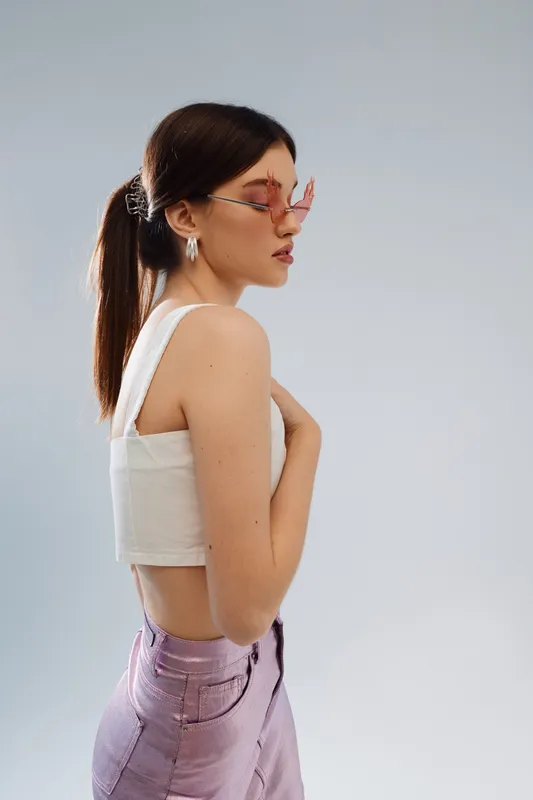 Young woman in stylish pink sunglasses and white crop top poses sideways against a light background. Concept of beauty, fashion, and modern style for lifestyle or portrait.