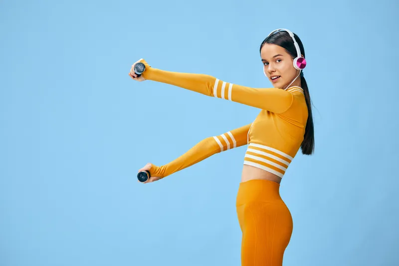 Woman fitness enthusiast performs a dynamic arm workout with dumbbells in a bright blue studio. Athletic outfit in orange, headphones on, focused expression, energetic movement showcasing strength