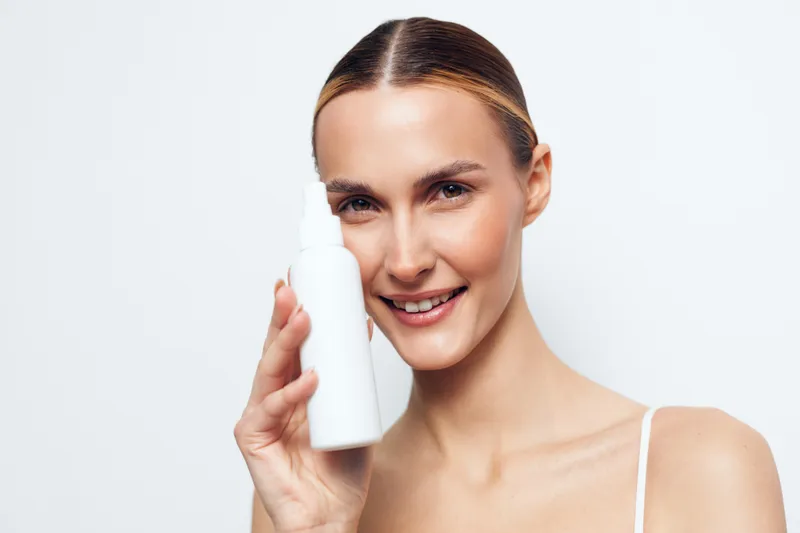 Smiling woman with clear skin holding a white cosmetic bottle close to her face on a white background. Concept of skincare, beauty, health, and personal care with natural makeup.