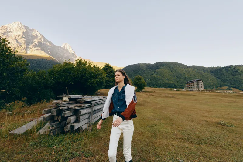 Woman walking in a mountain meadow, nature landscape with rustic wooden beams and distant abandoned building. Casual outfit, serene outdoor scene and countryside exploration.