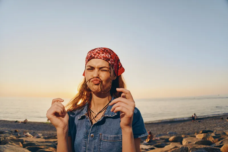 playful woman making funny face by the sea wearing red bandana and denim shirt during golden hour sunset on rocky beach