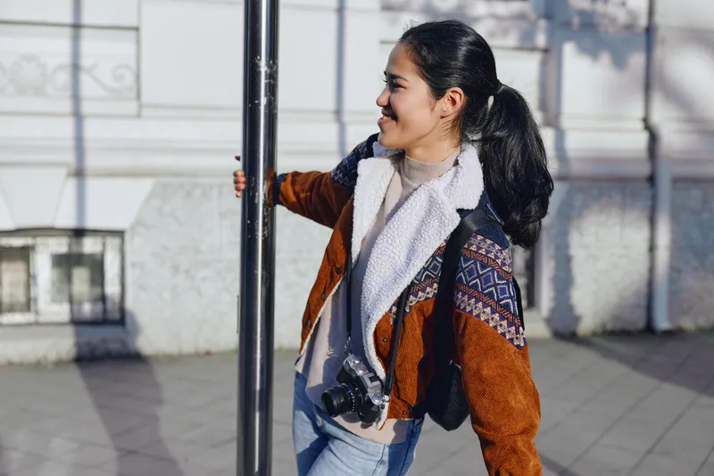 Camera in hand, stylish traveler strolls outdoors along a street, wearing a warm brown jacket and patterned scarf, capturing the moment with a casual candid vibe under sunlight.