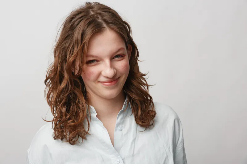 Smiling young woman with curly brown hair wearing a white shirt looks confidently at the camera on a plain light background. Concept of natural beauty and casual portrait.