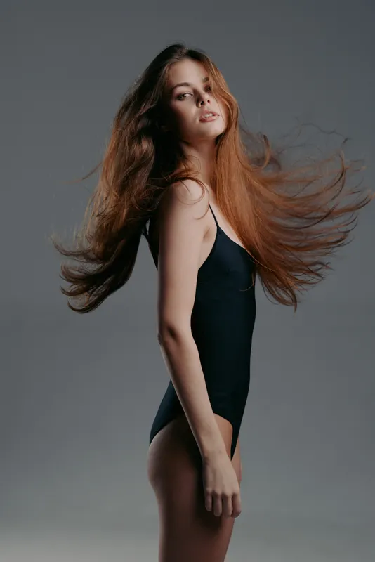 Young woman with flowing hair in black bodysuit posing against gray background, beauty and elegance concept