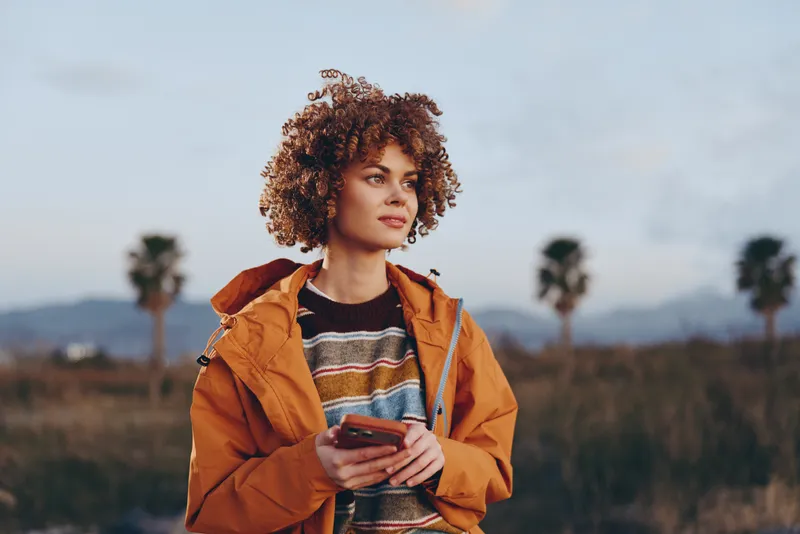Woman lifestyle outdoors, curly hair, orange jacket, holding smartphone, confident look, standing in grassy field, evening sky, travel mood, palm trees far away
