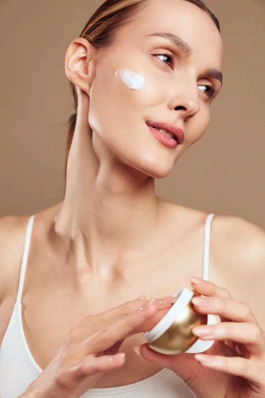 Young woman with blonde hair applies moisturizing cream on her cheek while holding a jar in a skincare routine against a neutral background.