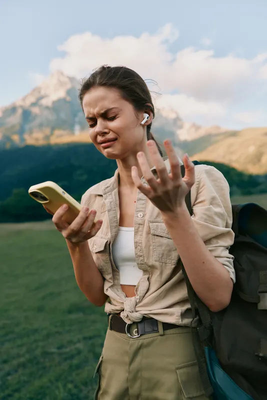A woman enjoys outdoor lifestyle, checking her phone while wearing earbuds and carrying a backpack, on a casual hike in a scenic landscape with mountains and lake in the background.