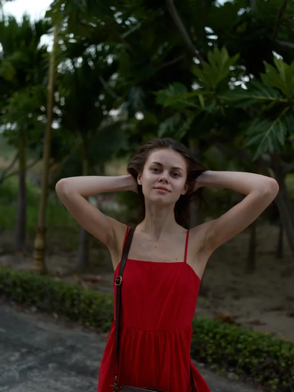 Young woman in red dress standing outdoors with hands in hair, surrounded by tropical greenery and soft natural light.