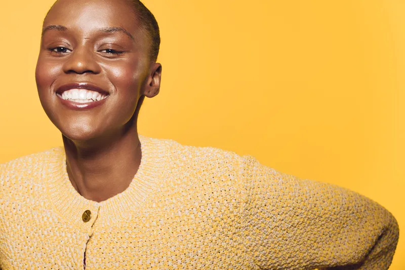 Smiling black African woman with dark skin wearing yellow knitted sweater poses against yellow background. Concept of happiness, fashion, and natural beauty with warm studio lighting.