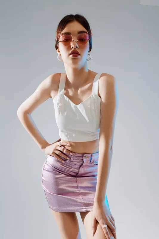 Young woman in a white top and shiny pink skirt poses confidently wearing pink sunglasses. Studio shot with soft lighting. Concept of beauty fashion style and youth.