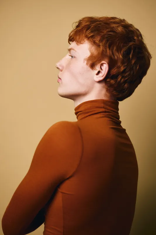 Profile of young man with red hair wearing brown turtleneck sweater against plain beige background. Side view, minimalistic, casual style portrait with clean composition and soft lighting.