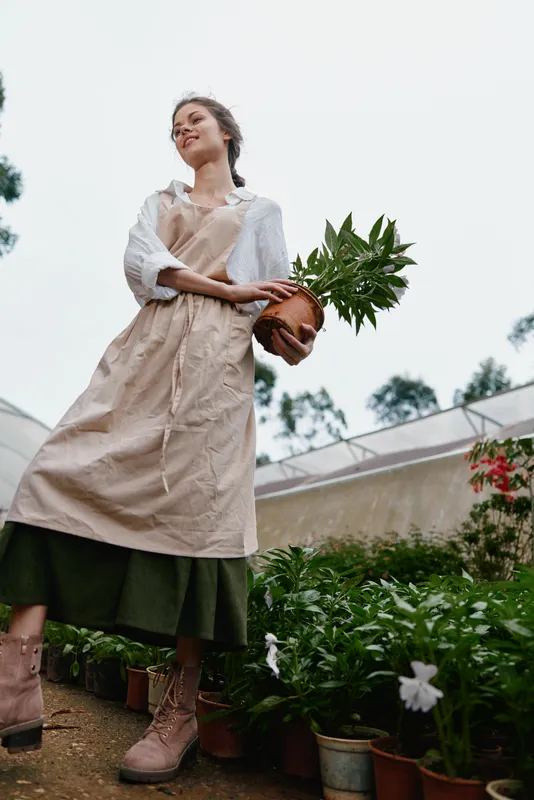 Young woman in a garden wearing an apron, smiling while holding a potted plant, surrounded by greenery, showcasing a sustainable lifestyle in a natural setting