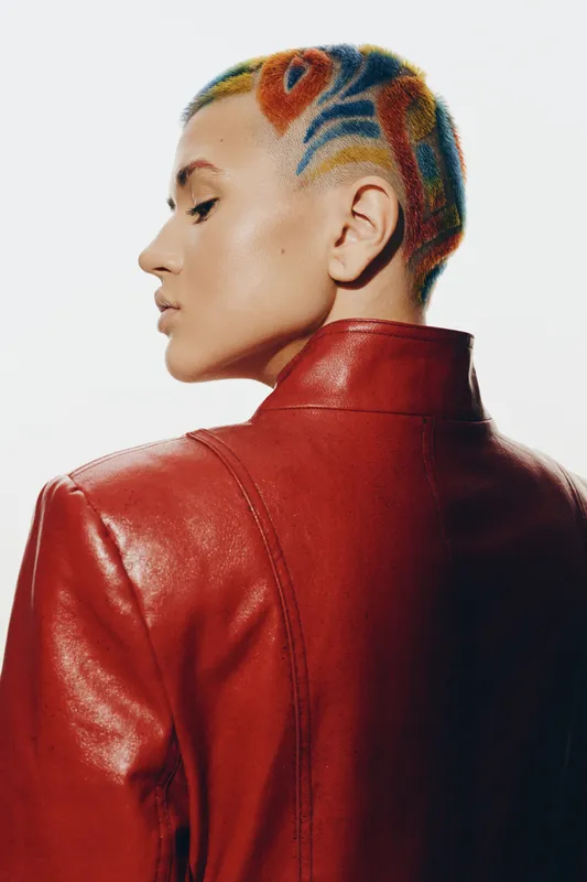 woman with colorful painted hair wearing a red leather jacket, side profile with a neutral expression, creative hairstyle on light background, modern fashion look.