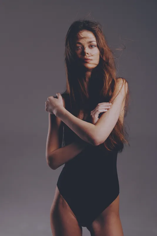 young woman in black bodysuit posing with arms crossed against gray background with soft lighting