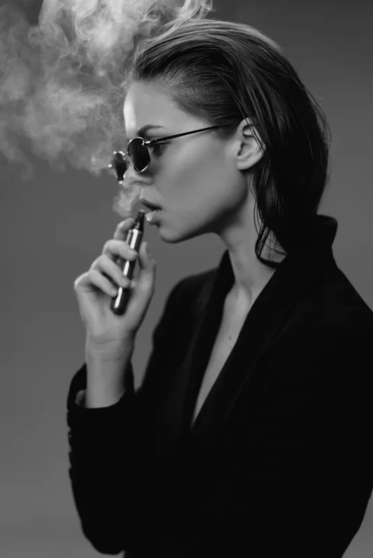 Black and white portrait of a woman vaping with sunglasses, smoke, and stylish dark clothing