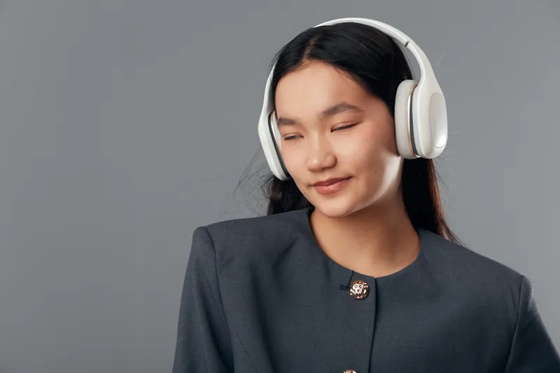 Young woman with long dark hair wearing white headphones listening to music with eyes closed, smiling slightly against plain gray background in studio portrait.