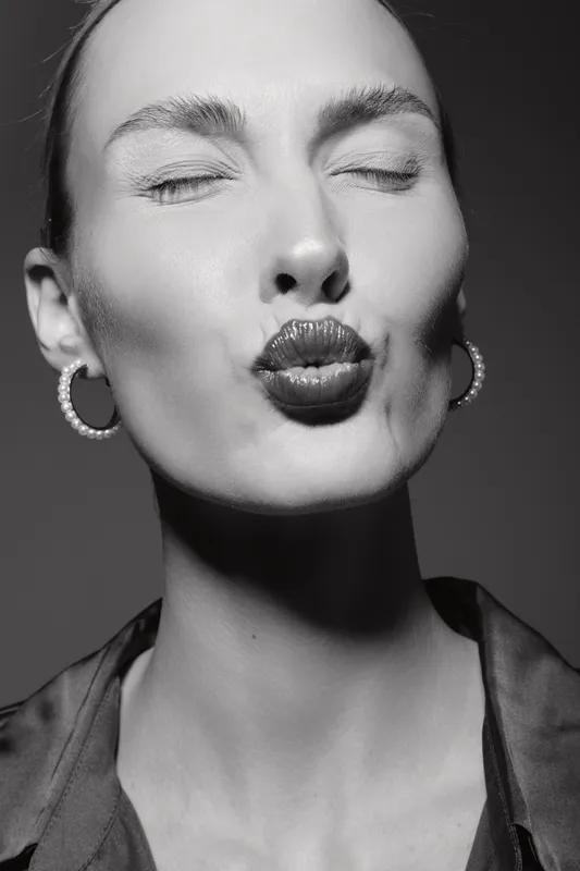 Caucasian woman with pale skin and closed eyes sends a kiss, wearing hoop earrings and silky blouse in black and white.