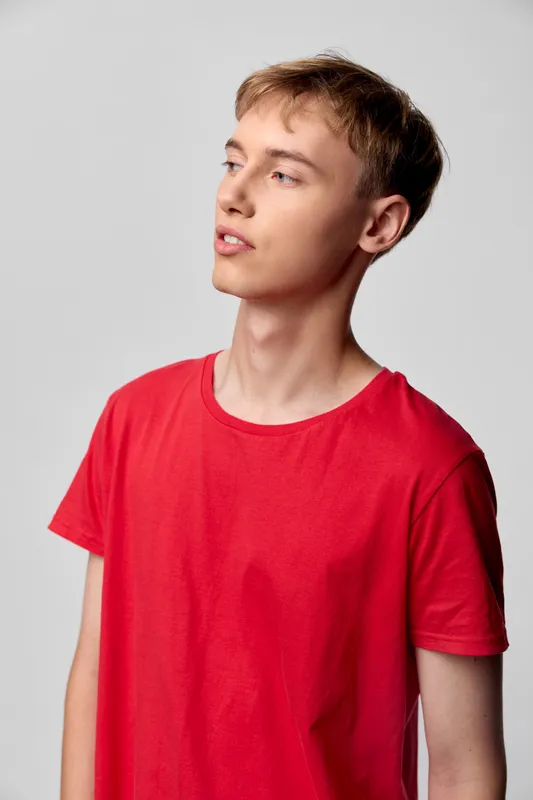 Young man wearing a red shirt stands against a neutral studio backdrop, looking off to the side with a calm gaze, capturing casual fashion and modern minimalism in a clean, commercial style suitable