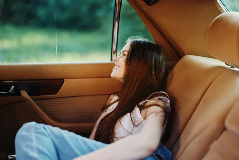 Young woman enjoying a carefree moment in a vintage car, smiling with long hair against a green outdoor backdrop on a sunny day.