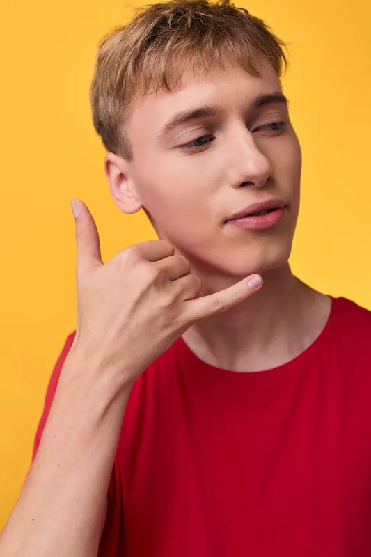 Hand gesture, call me concept, yellow background, red shirt, young man. Casual studio shot conveying communication, invitation, or friendly outreach through a simple signaling pose and relaxed