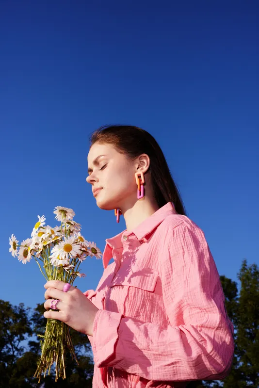 Young woman in a pink shirt holding daisies, enjoying a sunny day outdoors, with a clear blue sky in the background, radiating joy and confidence