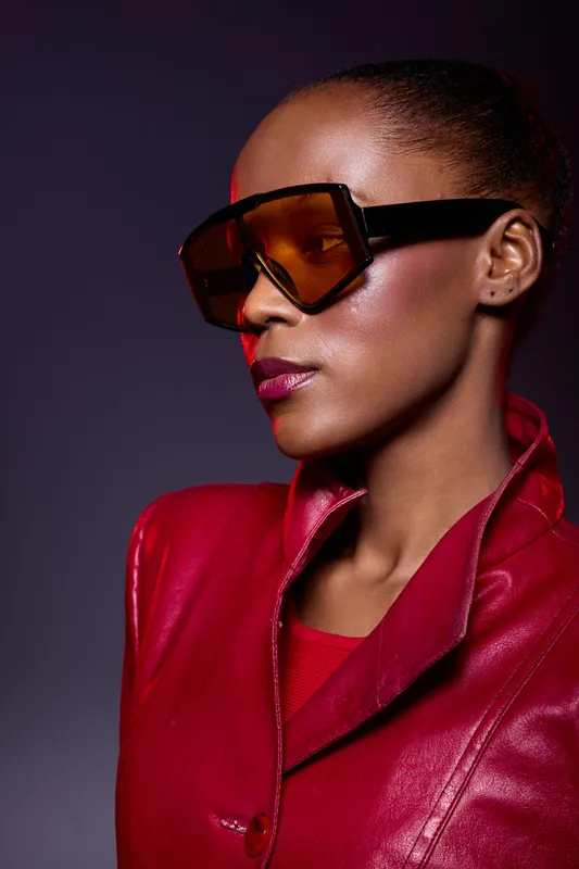 Stylish African American woman with dark skin wearing large sunglasses and a red leather jacket looks confident in a studio with dramatic lighting. Fashion and elegance concept.