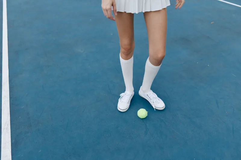 Female tennis player in a white skirt and knee high socks standing on a blue tennis court with a yellow tennis ball, exuding confidence and energy