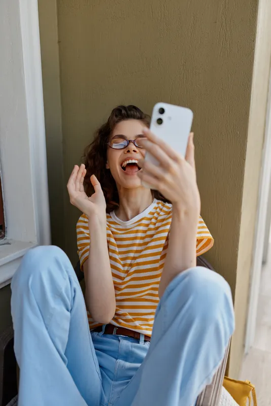 Smiling woman with striped shirt taking a selfie indoors, casual home setting with light walls, warm mood, playful energy, relaxed pose, modern lifestyle.
