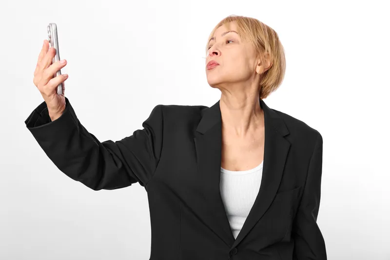 Woman smartphone selfie mature business suit portrait caucasian senior using front camera, confident social media moment, studio shot isolated minimal background