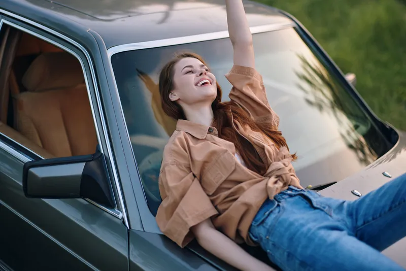 Cheerful young woman enjoying a sunny day, lounging on a vintage car in casual attire, expressing happiness in a vibrant green outdoor setting.