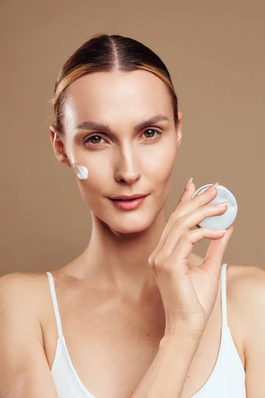 Woman skincare portrait, young adult holds cotton pad, applies cream on face, fresh clean skin, spa hygiene concept, beige background, studio lighting