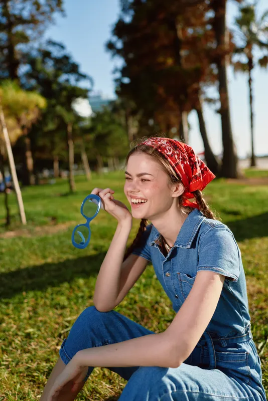 Lifestyle woman in modern boho-western denim street style sitting outdoors smiling with blue sunglasses on a sunny day, film color effect enhancing warm tones.