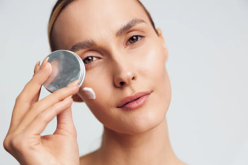 A close-up of a young woman applying makeup with a compact powder, soft lighting highlights smooth skin, conveying a calm beauty routine and self-care moment.