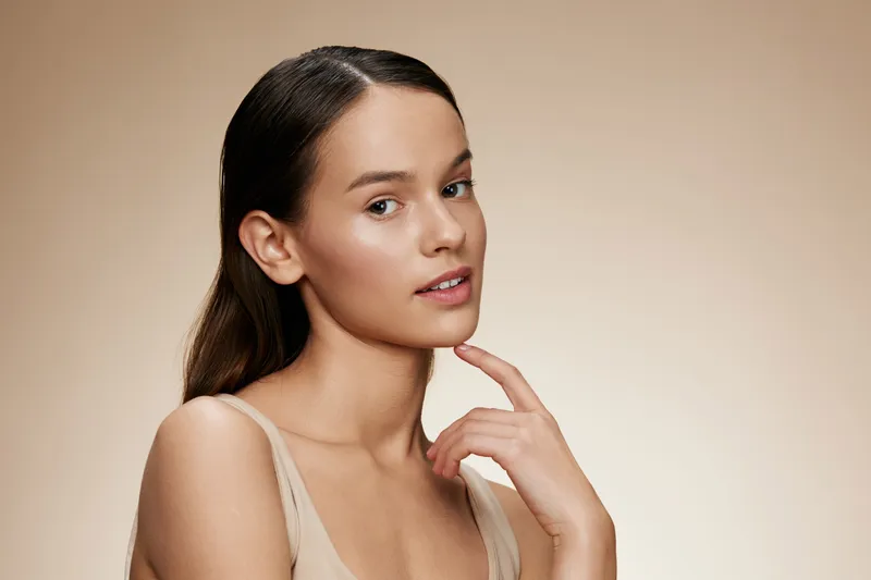 Natural beauty portrait of a young woman with smooth skin, wearing minimal makeup, showcasing a serene expression against a soft beige background