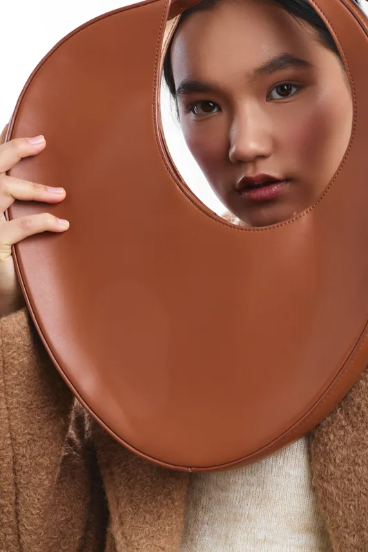 Asian woman peers through a circular leather frame, fashion forward pose in warm tones, studio lighting highlights texture and contemporary elegance, and confident expression.