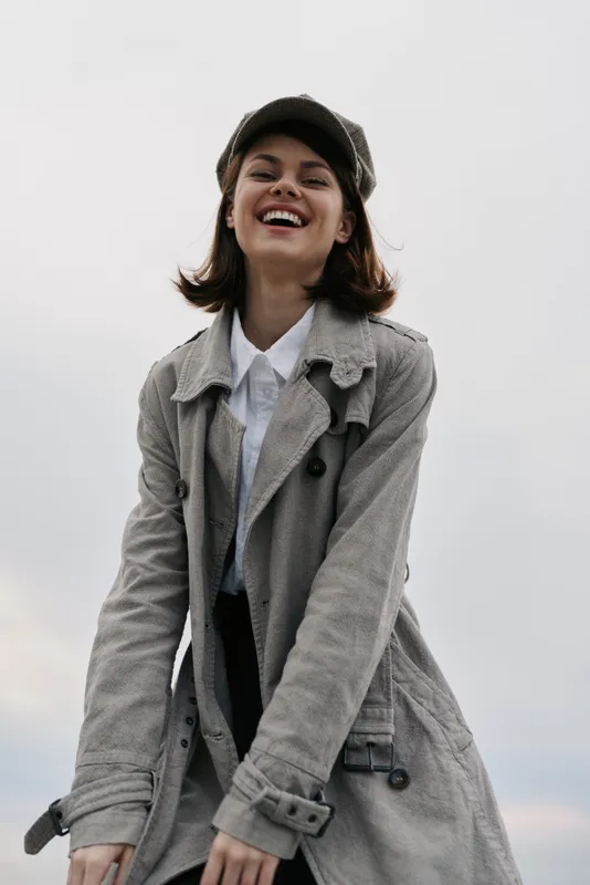 Happy young woman wearing a grey coat and matching hat smiles outdoors against a cloudy sky. Casual fashion, autumn style, joyful mood, and natural beauty concept.