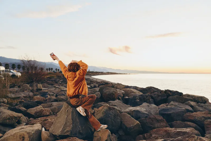 Woman lifestyle outdoors among rocks on the sea at sunset, arms raised in a stretch, wearing a warm jacket, enjoying nature, exercise and calm golden light, quiet evening mood