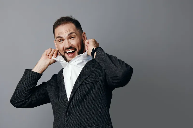 A confident business professional man in a textured blazer pulls up his white shirt with a surprised grin, showcasing decisive energy against a solid gray background.