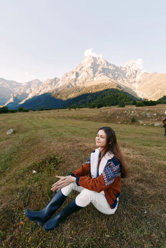 woman mountains meadow sweater boots nature portrait relaxation: young woman sits on grassy meadow wearing a cozy knit sweater and rubber boots, smiling peacefully while enjoying alpine mountain