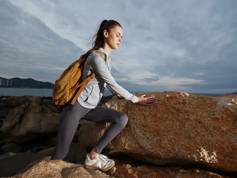 Young woman practicing yoga outdoors, wearing athletic clothing with a backpack, showing determination against a natural landscape backdrop